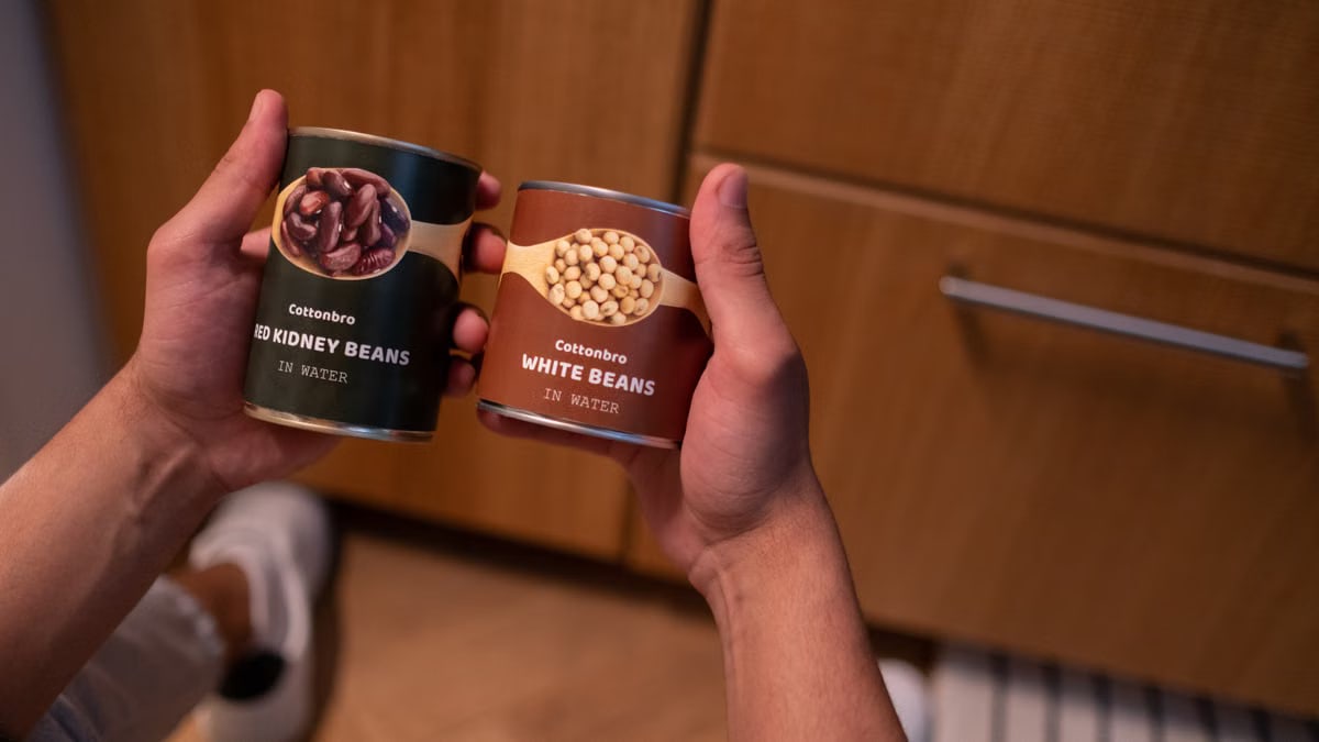 A person holding canned red kidney beans and white beans in a kitchen setting, emphasizing food storage.